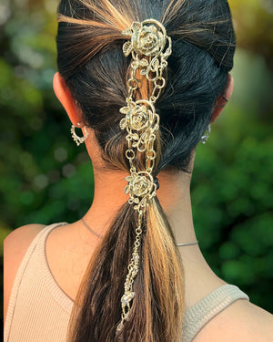 Gold Rose Hair Armor ponytail holder worn by model, featuring ornate rose medallions, cascading chains, and crystal accents securing a ponytail, rear view against a blurred outdoor background.