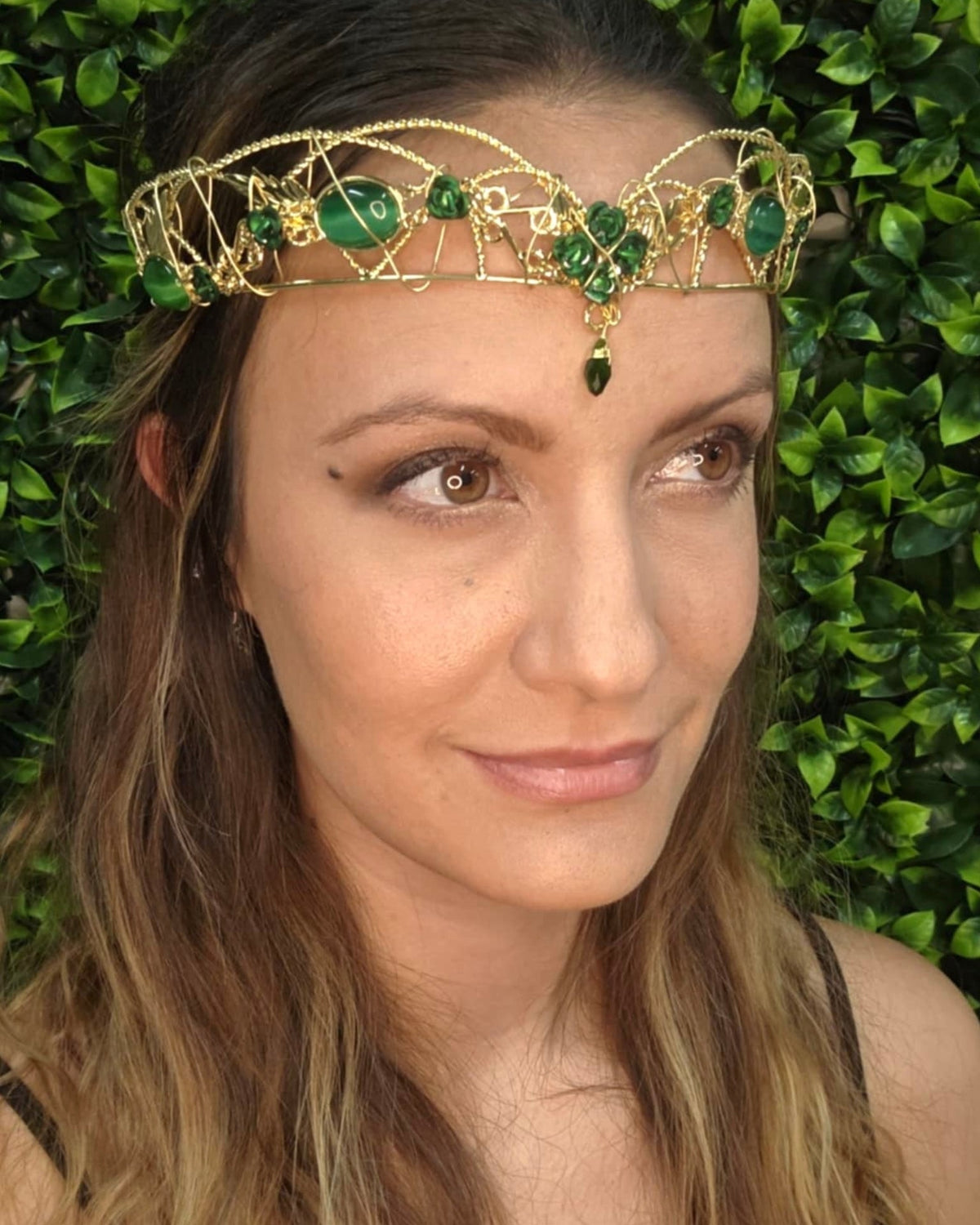 Model wearing a gold fantasy crown headpiece with green gemstones and floral wire design