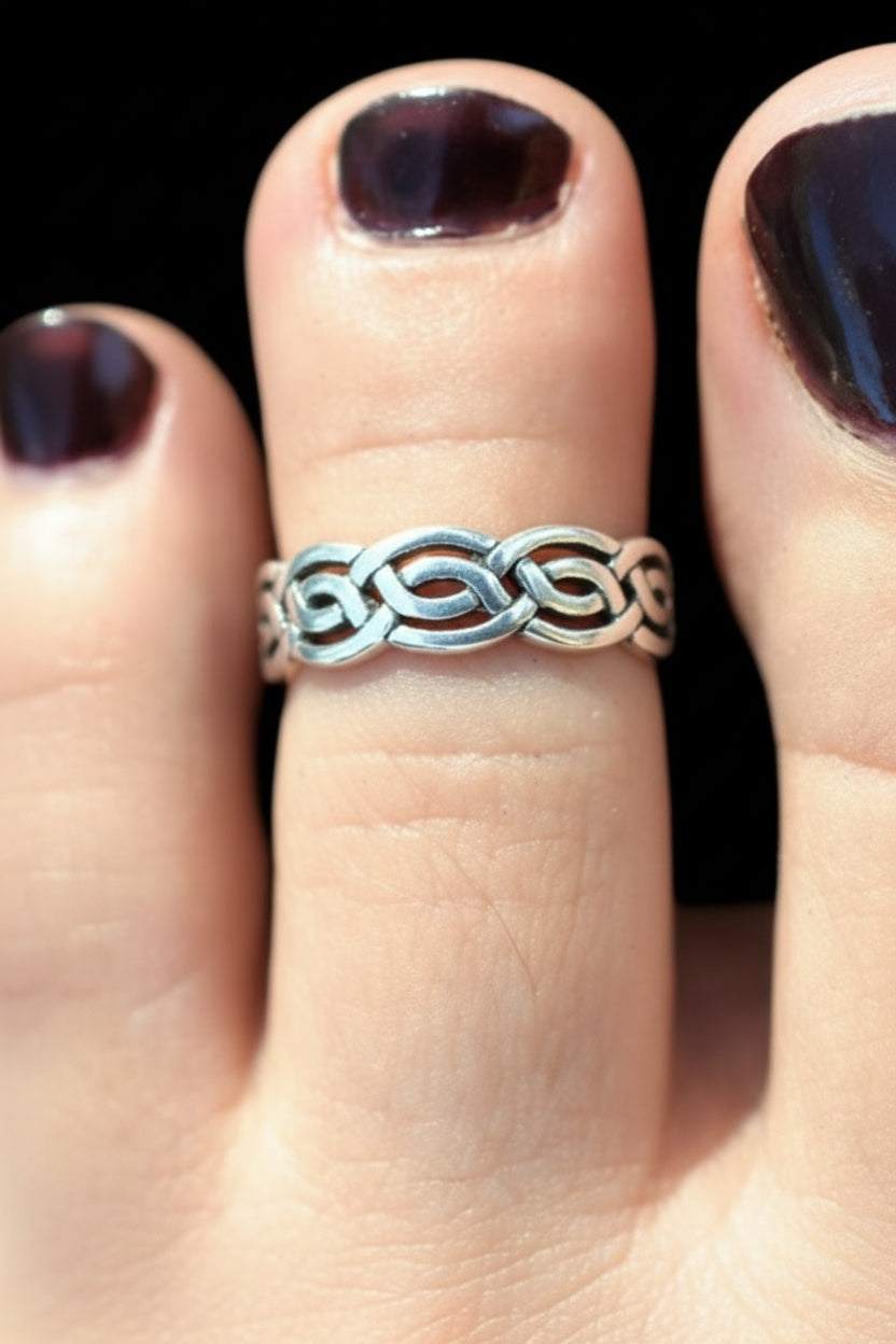 A close-up of a person's toes wearing a silver-toned interlocking knot ring on the middle toe, with dark brown nail polish against a black background.